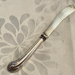 K&ouml;nnte beinhalten: Ein silbernes Buttermesser mit gebogener Klinge und dekorativem Griff. Der Griff hat ein abgerundetes, verziertes Design am Fu&szlig;. Das Messer liegt auf einem hellen Hintergrund mit floralem Muster.