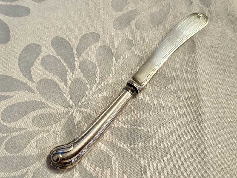 K&ouml;nnte beinhalten: Ein silbernes Buttermesser mit gebogener Klinge und dekorativem Griff. Der Griff hat ein abgerundetes, verziertes Design am Fu&szlig;. Das Messer liegt auf einem hellen Hintergrund mit floralem Muster.