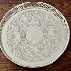 May include: A round silver-toned serving tray featuring an intricate engraved floral and scrollwork pattern across the center surface. The tray has a raised, openwork gallery rim with a repeating geometric lattice design along the edge.