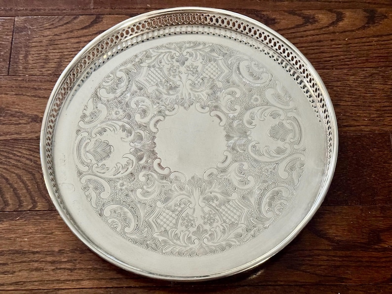 May include: A round silver-toned serving tray featuring an intricate engraved floral and scrollwork pattern across the center surface. The tray has a raised, openwork gallery rim with a repeating geometric lattice design along the edge.