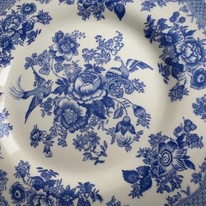 K&ouml;nnte beinhalten: Ein wei&szlig;er Keramikteller mit einem blauen und wei&szlig;en Blumenmuster. Das Muster zeigt einen stilisierten Vogel, Blumen und Bl&auml;tter. Der Teller hat einen gewellten Rand.