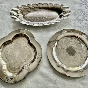 May include: Three antique silver serving dishes. The top dish is oval with a scalloped edge. The bottom left dish is quatrefoil-shaped. The bottom right dish is round with a decorative edge. All dishes have ornate engraved patterns.