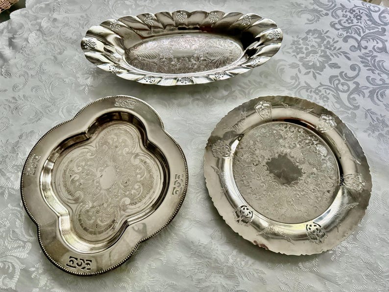 May include: Three antique silver serving dishes. The top dish is oval with a scalloped edge. The bottom left dish is quatrefoil-shaped. The bottom right dish is round with a decorative edge. All dishes have ornate engraved patterns.