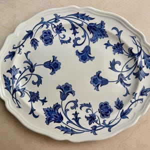 May include: An oval ceramic serving platter with a scalloped edge, featuring a white base color and a decorative pattern of dark blue floral vines and leaves that curve along the perimeter and extend toward the center of the dish.