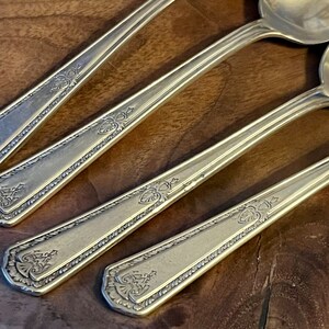 Pu&ograve; includere: Primo piano di quattro cucchiai d'argento con decorazioni floreali e a foglie ornate sui manici. I cucchiai hanno una superficie lucida e sono disposti in diagonale. I cucchiai sono su uno sfondo di legno scuro, evidenziando l'argenteria.