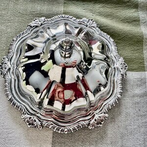 May include: Ornate silver serving dish with a central handle and multiple compartments. The dish has a decorative, scalloped edge and a reflective surface, perfect for serving appetizers or small portions.