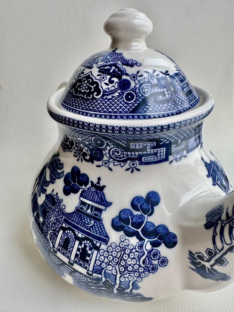 K&ouml;nnte beinhalten: Blaue und wei&szlig;e Porzellan-Teekanne mit traditionellem chinesischem Weidenmuster. Die Teekanne hat einen runden Korpus mit Deckel und Ausguss. Das Muster zeigt eine Pagode, B&auml;ume und Blumen.