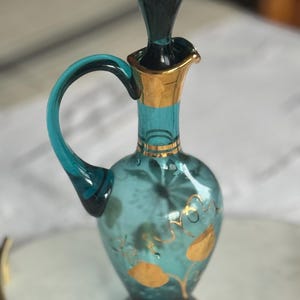 May include: A teal blue glass bottle with a gold rim and floral design. The bottle has a stopper and a handle.