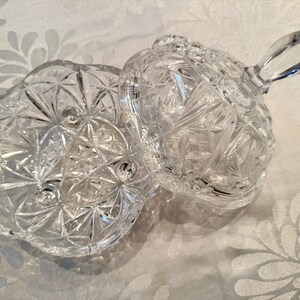May include: A clear, pressed glass candy dish with a matching lid. The dish features a faceted starburst pattern, a scalloped rim, and three small ball feet. The lid includes a pointed, vertical handle, and the entire piece is made of transparent glass.