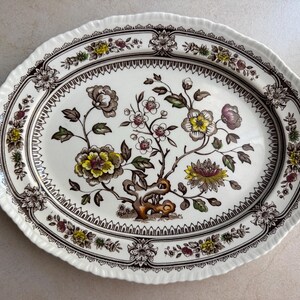 May include: An oval ceramic serving platter with a cream-colored base and a scalloped edge. The center features a detailed botanical illustration of flowers in shades of yellow, brown, and green, surrounded by a dark brown decorative border.
