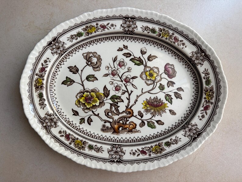 May include: An oval ceramic serving platter with a cream-colored base and a scalloped edge. The center features a detailed botanical illustration of flowers in shades of yellow, brown, and green, surrounded by a dark brown decorative border.