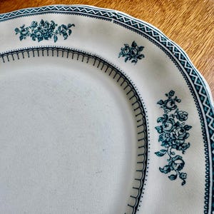 May include: A cream-coloured ceramic plate with a scalloped edge featuring a dark teal floral pattern and a geometric border. The design includes small clusters of roses and leaves arranged around the rim, complemented by a thin line of arches.