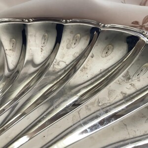 K&ouml;nnte beinhalten: Eine silberfarbene Metallschale in Form einer Jakobsmuschel mit einer polierten, reflektierenden Oberfl&auml;che. Die Schale weist tiefe, strahlenf&ouml;rmige Rillen auf, die von der Mitte zum gewellten Rand verlaufen und ein f&auml;cherartiges Muster bilden.