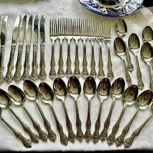 May include: An assortment of vintage silverware, including knives, forks, and spoons, arranged on a white tablecloth. The silverware has ornate handles and a reflective, silver finish. A blue and white patterned plate is visible in the background.