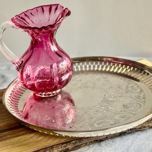 Puede incluir: Una jarra de cristal de color rub&iacute; con asa transparente y borde ondulado se encuentra sobre una bandeja plateada con un dise&ntilde;o floral ornamentado. La bandeja descansa sobre una tabla de madera.