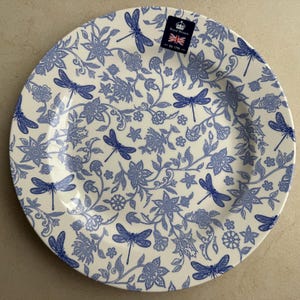 Puede incluir: Un plato redondo de cer&aacute;mica con una base de color crema y un patr&oacute;n azul repetitivo de lib&eacute;lulas, flores y enredaderas. Una peque&ntilde;a etiqueta rectangular en la parte superior muestra un icono de corona, el texto Royal Winton y una bandera brit&aacute;nica.