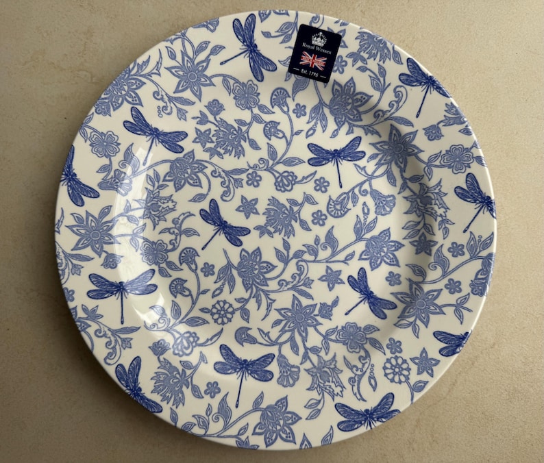 Puede incluir: Un plato redondo de cer&aacute;mica con una base de color crema y un patr&oacute;n azul repetitivo de lib&eacute;lulas, flores y enredaderas. Una peque&ntilde;a etiqueta rectangular en la parte superior muestra un icono de corona, el texto Royal Winton y una bandera brit&aacute;nica.