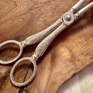 May include: Silver-toned ornate serving tongs with a floral design. The tongs are on a wooden surface.