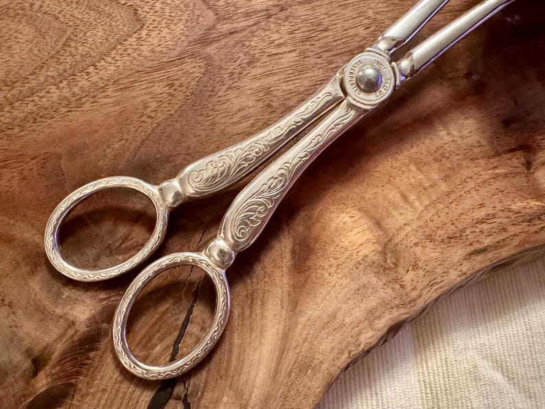 May include: Silver-toned ornate serving tongs with a floral design. The tongs are on a wooden surface.