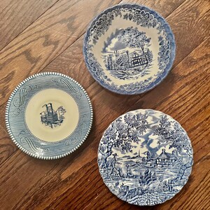 May include: A collection of three ceramic pieces featuring blue and white transferware patterns. One bowl displays a landscape with a house, while two plates feature a steamboat illustration and a rural scene with a person and a dog respectively.