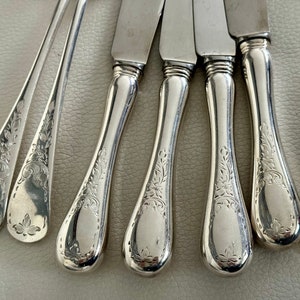 May include: Close-up of a collection of silver cutlery, including knives and spoons. The handles feature an ornate floral design. The silverware is arranged on a light-colored surface, reflecting the light.