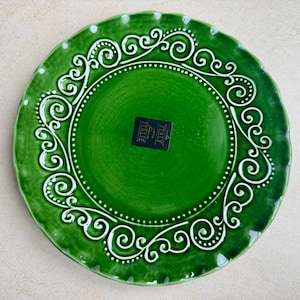 May include: A round, emerald green ceramic plate with a scalloped edge. The rim features a raised white scrollwork pattern and a dotted border. A black rectangular sticker in the center displays the text ITALY and ITALIE in white lettering.