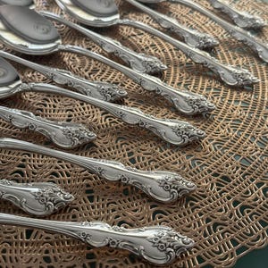 May include: Close-up of a collection of ornate silver spoons. The spoons have a reflective surface and detailed floral patterns on the handles. They are arranged on a textured, golden-brown placemat.
