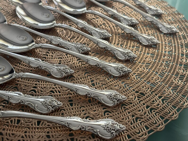 May include: Close-up of a collection of ornate silver spoons. The spoons have a reflective surface and detailed floral patterns on the handles. They are arranged on a textured, golden-brown placemat.