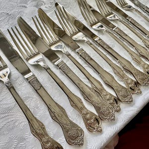 May include: An assortment of antique silverware, including forks and knives, arranged on a white tablecloth. The silverware has ornate floral detailing on the handles and a shiny, reflective surface. The image showcases a collection of vintage dining utensils.