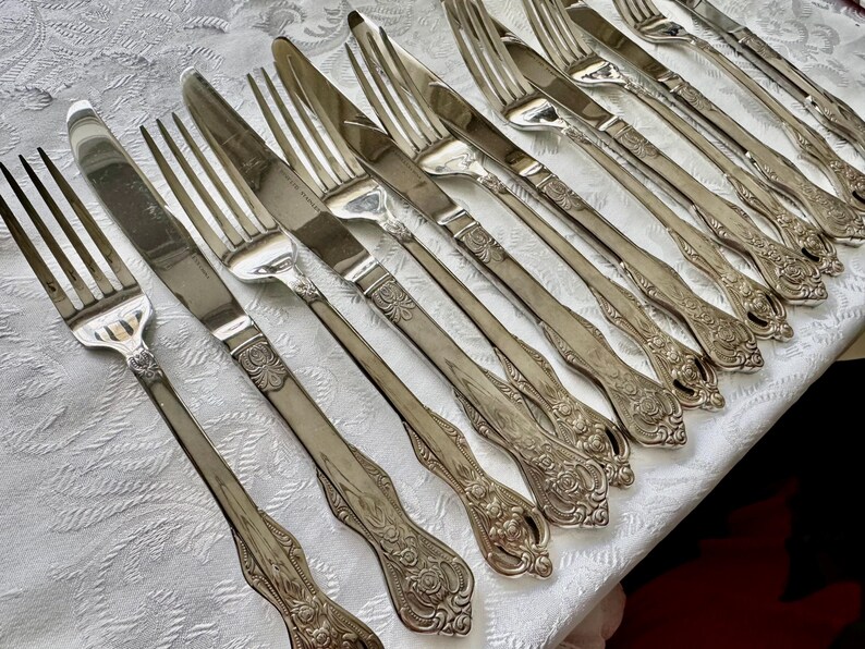 May include: An assortment of antique silverware, including forks and knives, arranged on a white tablecloth. The silverware has ornate floral detailing on the handles and a shiny, reflective surface. The image showcases a collection of vintage dining utensils.