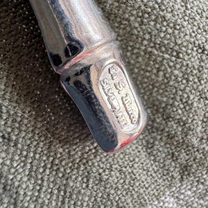 May include: Close-up of a silver-plated utensil with a rounded handle. The base of the utensil features an oval stamp with the text "Co. S. Marco Silverplate". The utensil is set against a textured, olive-green fabric.