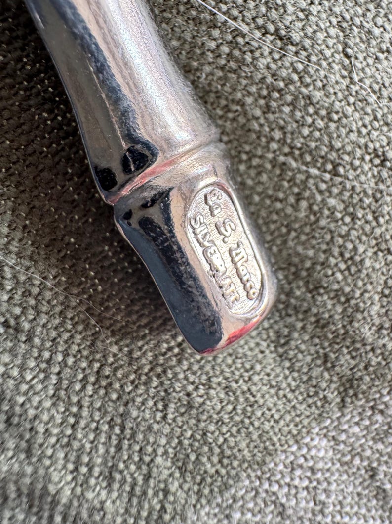 May include: Close-up of a silver-plated utensil with a rounded handle. The base of the utensil features an oval stamp with the text "Co. S. Marco Silverplate". The utensil is set against a textured, olive-green fabric.