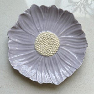 Puede incluir: Un plato de cer&aacute;mica con forma de flor y p&eacute;talos de color violeta claro que irradian desde un disco central. El centro presenta una superficie texturizada en relieve compuesta por numerosas cuentas peque&ntilde;as de color crema.