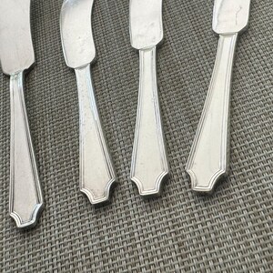 May include: Close-up of four silver butter knives with a decorative edge. The knives are arranged on a textured, grey surface. The blades are rectangular, and the handles have a tapered design.