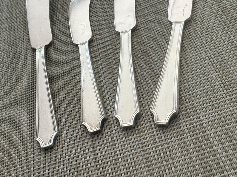 May include: Close-up of four silver butter knives with a decorative edge. The knives are arranged on a textured, grey surface. The blades are rectangular, and the handles have a tapered design.