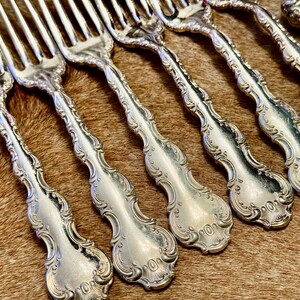 May include: A set of eight silver forks with ornate handles. The forks have a decorative design with a floral pattern. The forks are arranged in a row on a brown fur surface.