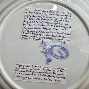 May include: The underside of a white ceramic dinner plate featuring a blue stamped maker's mark with the text AGLE inside an oval border. Two handwritten paper labels are attached to the surface, providing historical information about the plate.