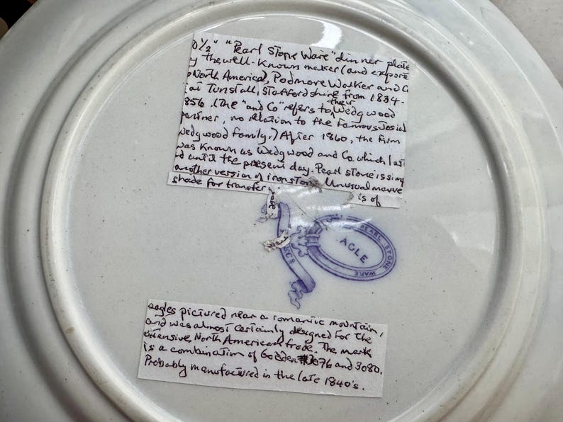 May include: The underside of a white ceramic dinner plate featuring a blue stamped maker's mark with the text AGLE inside an oval border. Two handwritten paper labels are attached to the surface, providing historical information about the plate.