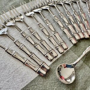 May include: A collection of silver-colored flatware, including forks and a ladle. The handles of the forks and ladle feature a bamboo design. The flatware is arranged on a patterned, neutral-toned tablecloth.