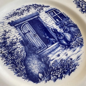 K&ouml;nnte beinhalten: Blaue und wei&szlig;e Keramikplatte mit einer Cottage-Szene mit einer T&uuml;r, einer Backsteinmauer und vier Kaninchen. Die Kaninchen stehen vor dem Cottage und sind von blauen Blumen umgeben.