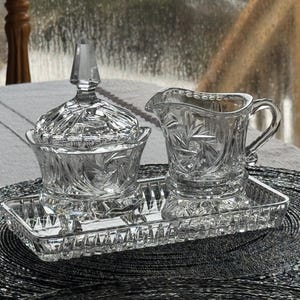 May include: A clear glass sugar bowl with a matching lid, a creamer pitcher, and a rectangular tray. Each piece features a pressed glass pattern with starburst and fan motifs. The sugar bowl has a pointed finial on the lid, and the creamer has a curved handle and a pouring spout.