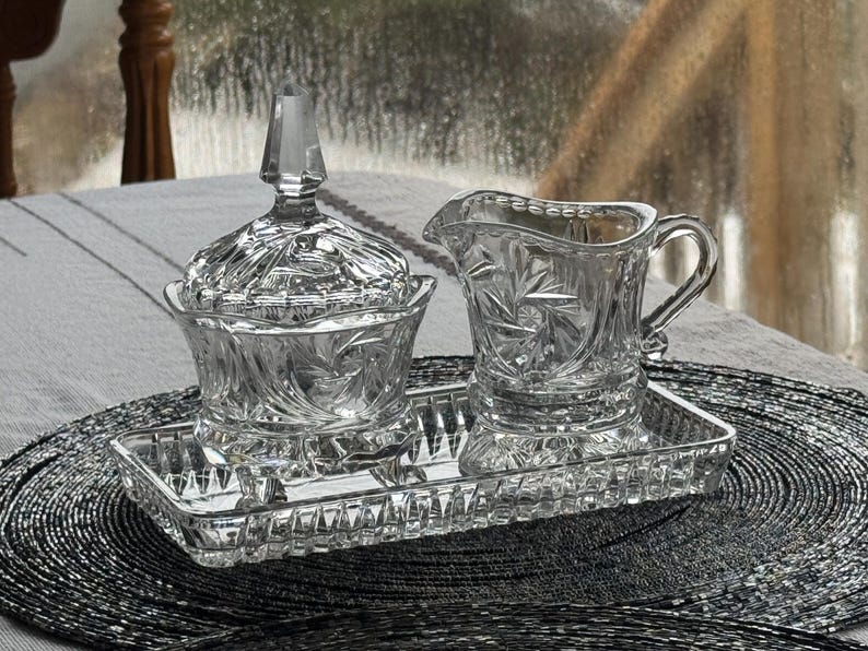 May include: A clear glass sugar bowl with a matching lid, a creamer pitcher, and a rectangular tray. Each piece features a pressed glass pattern with starburst and fan motifs. The sugar bowl has a pointed finial on the lid, and the creamer has a curved handle and a pouring spout.