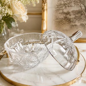 May include: A clear glass candy dish with a matching lid, featuring a faceted starburst pattern and a pointed handle. The bowl has a rounded shape with intricate geometric cuts throughout the glass surface, reflecting light across its transparent body.