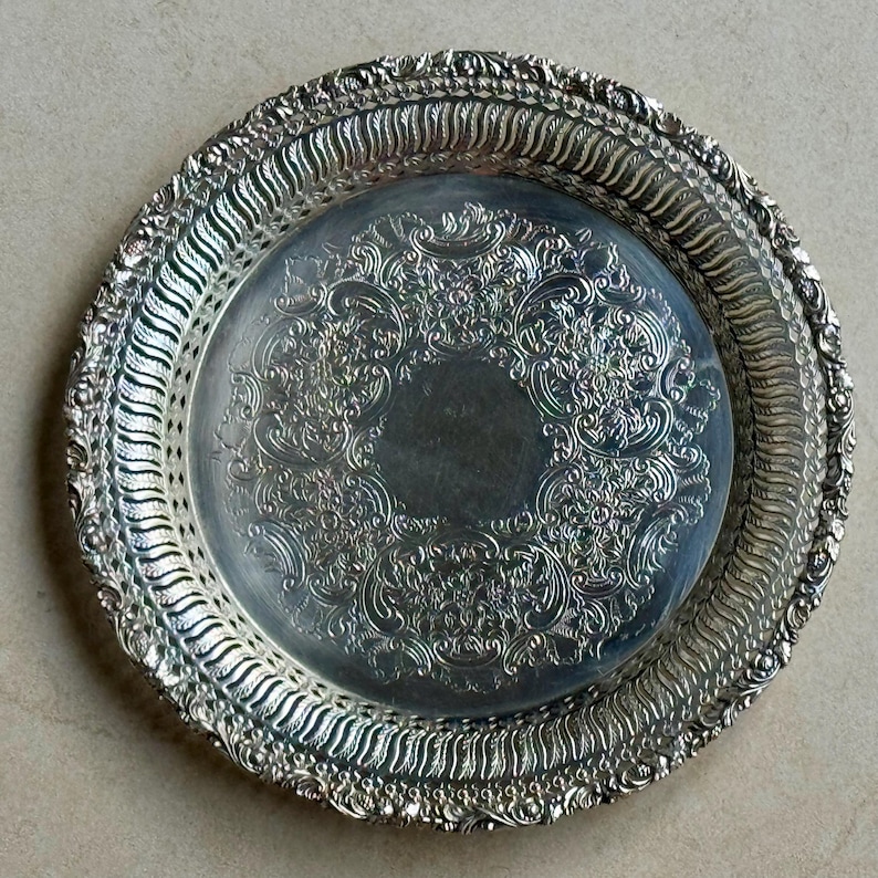 May include: A round silver-toned metal tray featuring an intricate embossed floral and scroll pattern in the center. The wide, raised rim has a decorative pierced lattice border with a scalloped edge and detailed relief work throughout the surface.