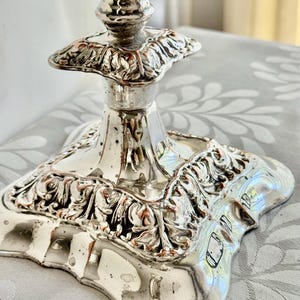 May include: A silver-toned lamp base featuring a square, ornate pedestal with intricate embossed leaf and scroll detailing. The metallic surface has a reflective finish with visible signs of wear and patina, showing a tapered neck design.