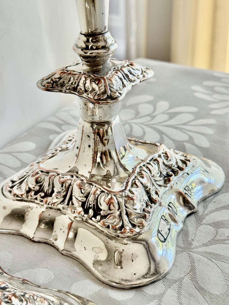 May include: A silver-toned lamp base featuring a square, ornate pedestal with intricate embossed leaf and scroll detailing. The metallic surface has a reflective finish with visible signs of wear and patina, showing a tapered neck design.