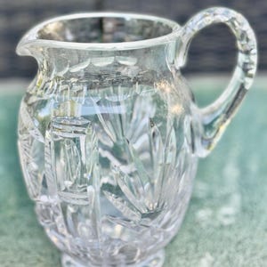 Puede incluir: Jarra de cristal transparente con cuerpo redondeado, pico vertedor y asa curva. La jarra presenta intrincados patrones de vidrio tallado, incluyendo l&iacute;neas verticales y formas de hojas. La base es un peque&ntilde;o pedestal circular.