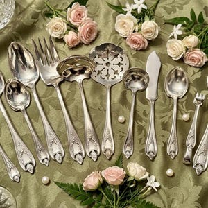 May include: A collection of silver-toned metal serving utensils featuring ornate embossed handles. The set includes various spoons, forks, a slotted server, and a flat-edged knife, all arranged on a green fabric surface with small pink roses.