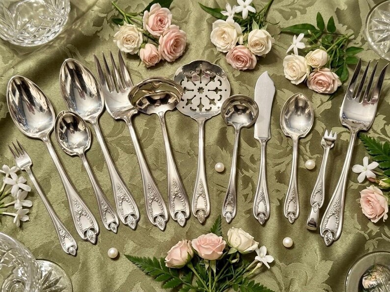 May include: A collection of silver-toned metal serving utensils featuring ornate embossed handles. The set includes various spoons, forks, a slotted server, and a flat-edged knife, all arranged on a green fabric surface with small pink roses.