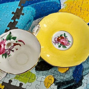 Pu&ograve; includere: Un set di tazza e piattino in porcellana composto da un piattino giallo brillante con un motivo floreale centrale e una tazza bianca decorata con un colorato bouquet di fiori rosa, bianchi e viola, entrambi rifiniti con un bordo dorato.